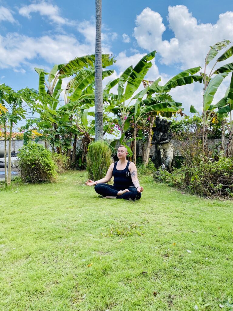 Photo of the owner of Accessible Align Yoga, Carolyn, sitting in meditation pose in a lush tropical garden with palm trees in the background.